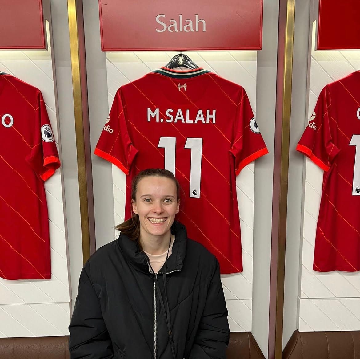 Maddie Hayes, Strength & Conditioning Coach at Carter & George Clinic in Hitchin, sitting in the Liverpool FC changing room in front of Mo Salah’s shirt.